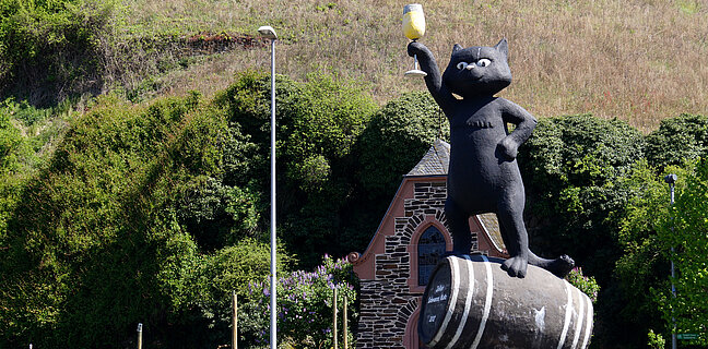 Skulptur einer schwarzen Katze, die auf einem Weinfass steht und ein Weinglas in die Höhe hält.