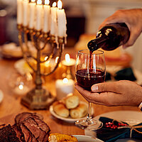 Im Vordergrund gießt jemand Rotwein in ein Glas, im Hintergrund ist ein Channukah-Leuchter und ein gedeckter Esstisch mit Kerzen zu sehen